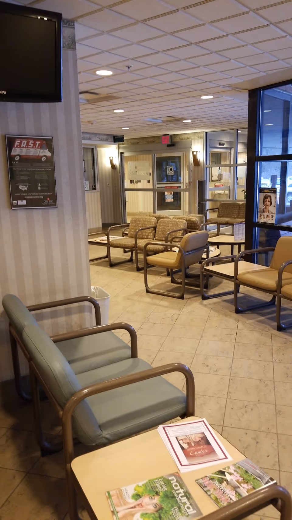 Waiting area in a hospital lobby with beige chairs, tables, magazines, and glass entrance doors.