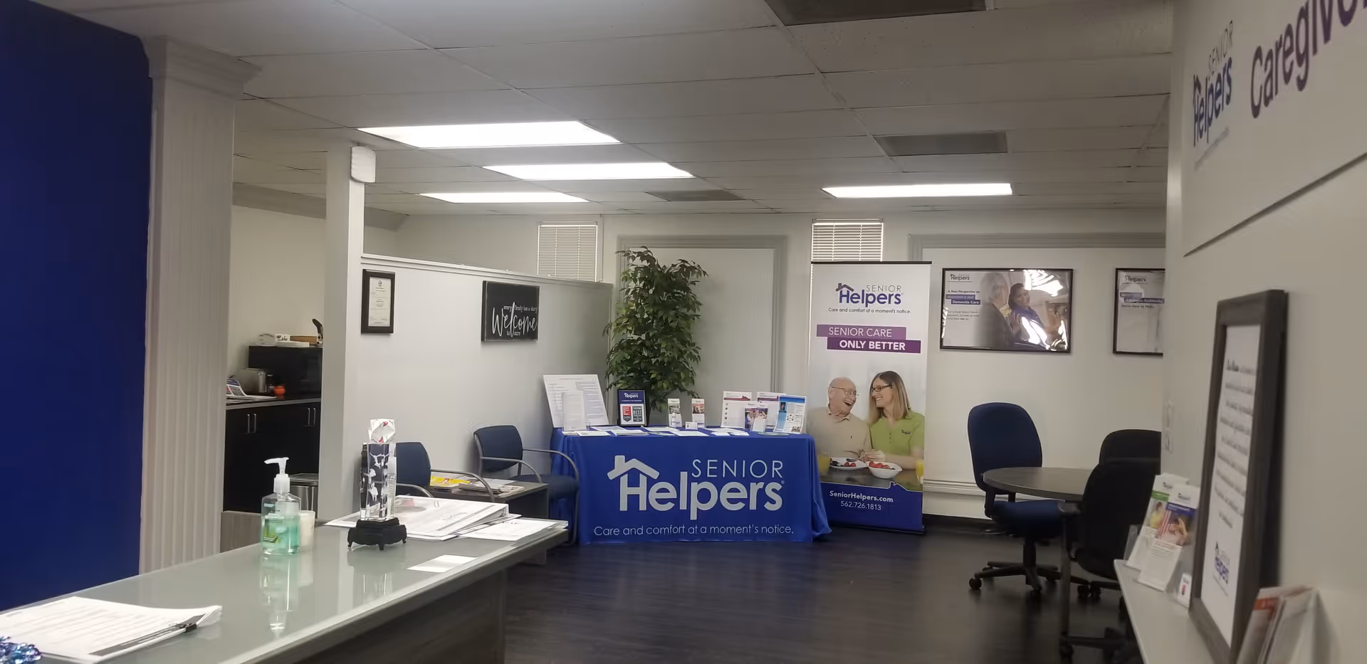 Lobby reception area with a desk, banners, chairs, and a plant in a Senior Helpers space.