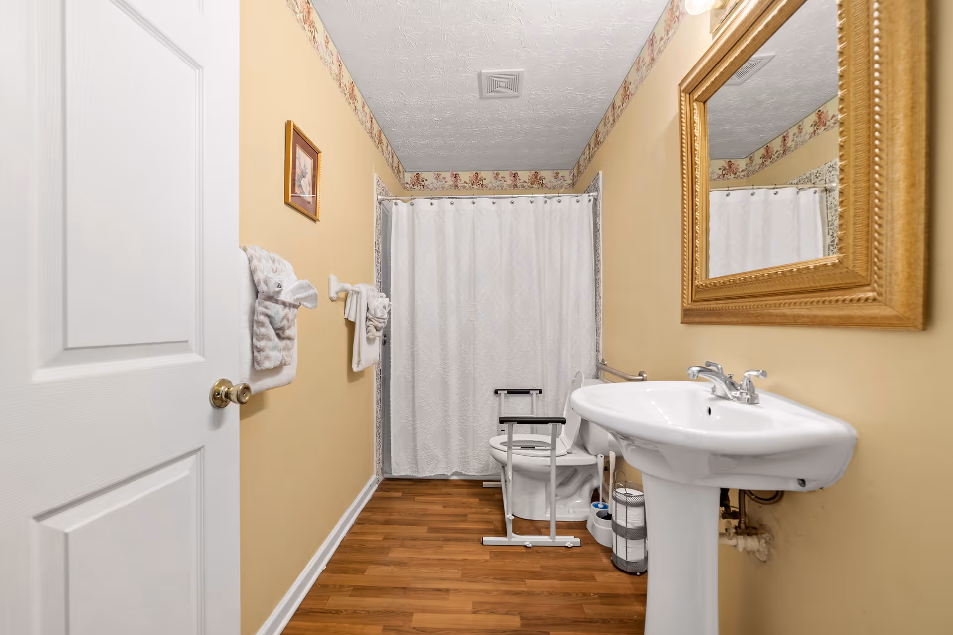 Small beige bathroom with pedestal sink, toilet, shower curtain, and a bathroom chair.