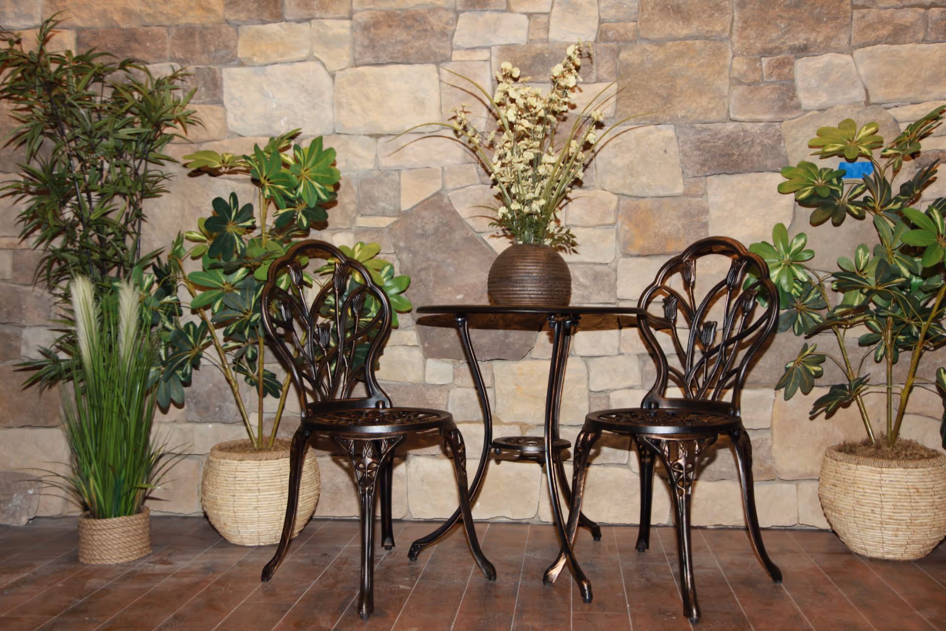Two dark metal chairs sit at a small table with a vase, against a stone wall.