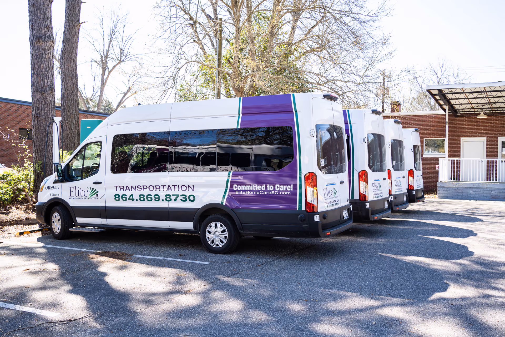 Row of Elite Home Care transportation vans parked in a lot beside a building.