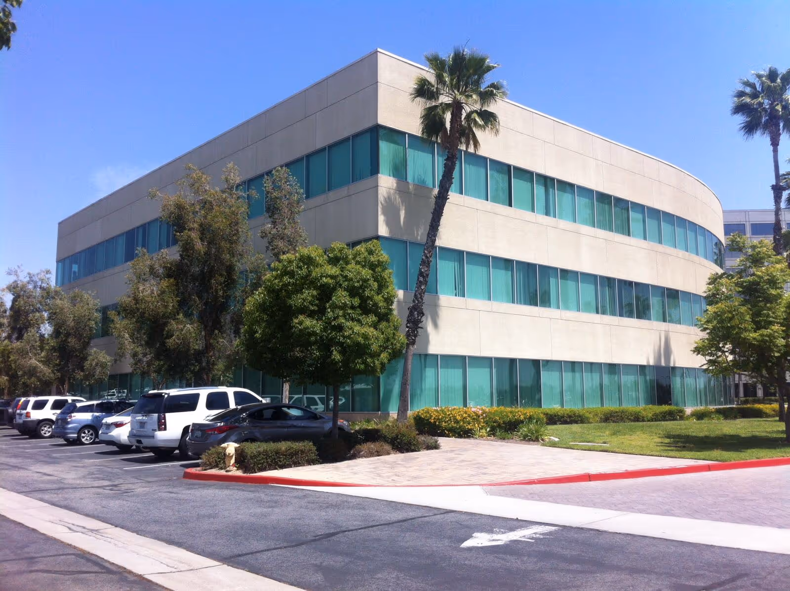 Modern beige office-style building with teal windows, palm trees, and a parking lot.