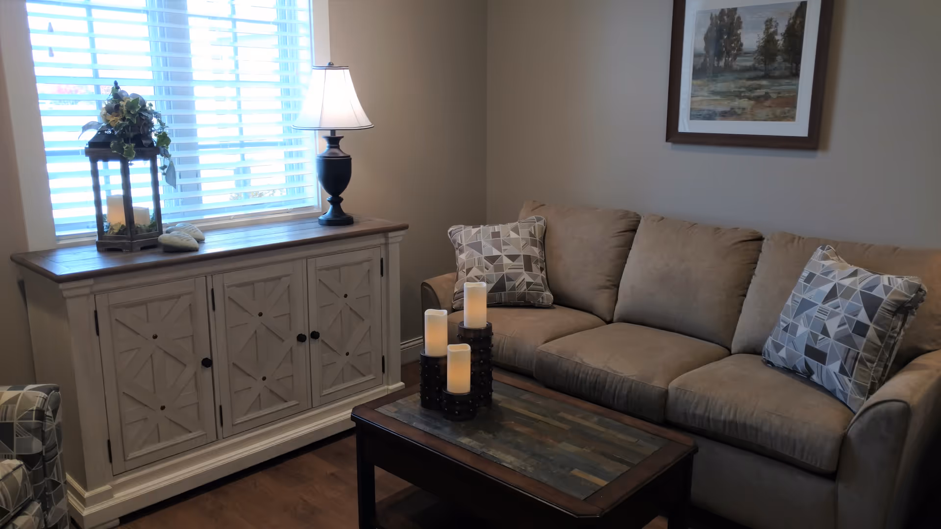 Cozy living room with beige sofa, coffee table with candles, cabinet, lamp, blinds, and art.
