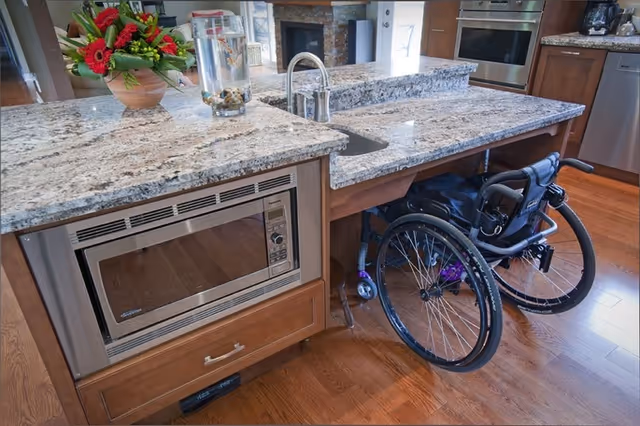 Wheelchair beside a kitchen island with granite countertops and a built-in microwave.