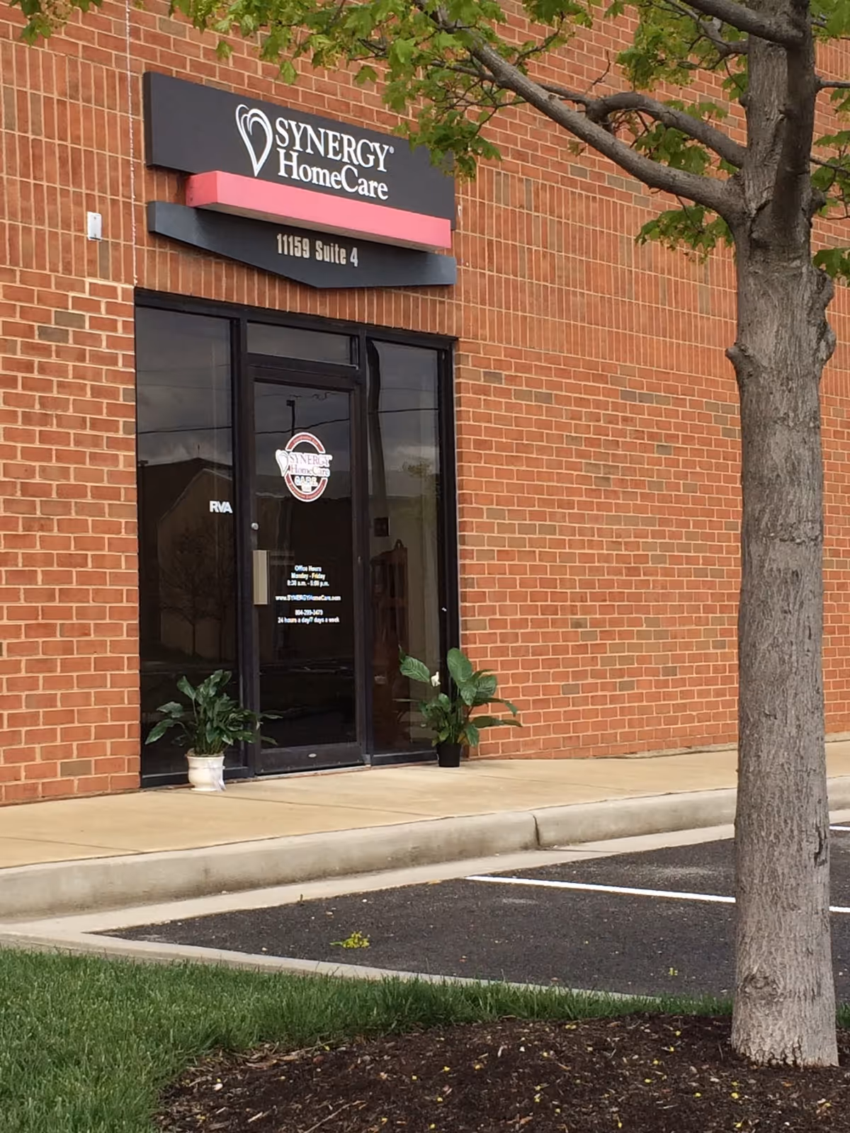 Brick building exterior with a Synergy HomeCare sign above a glass door and potted plants