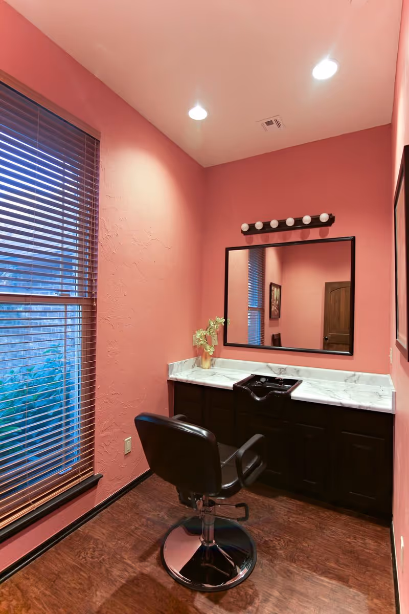 Pink-walled hair salon station with marble countertop, mirror, and black styling chair by a window.