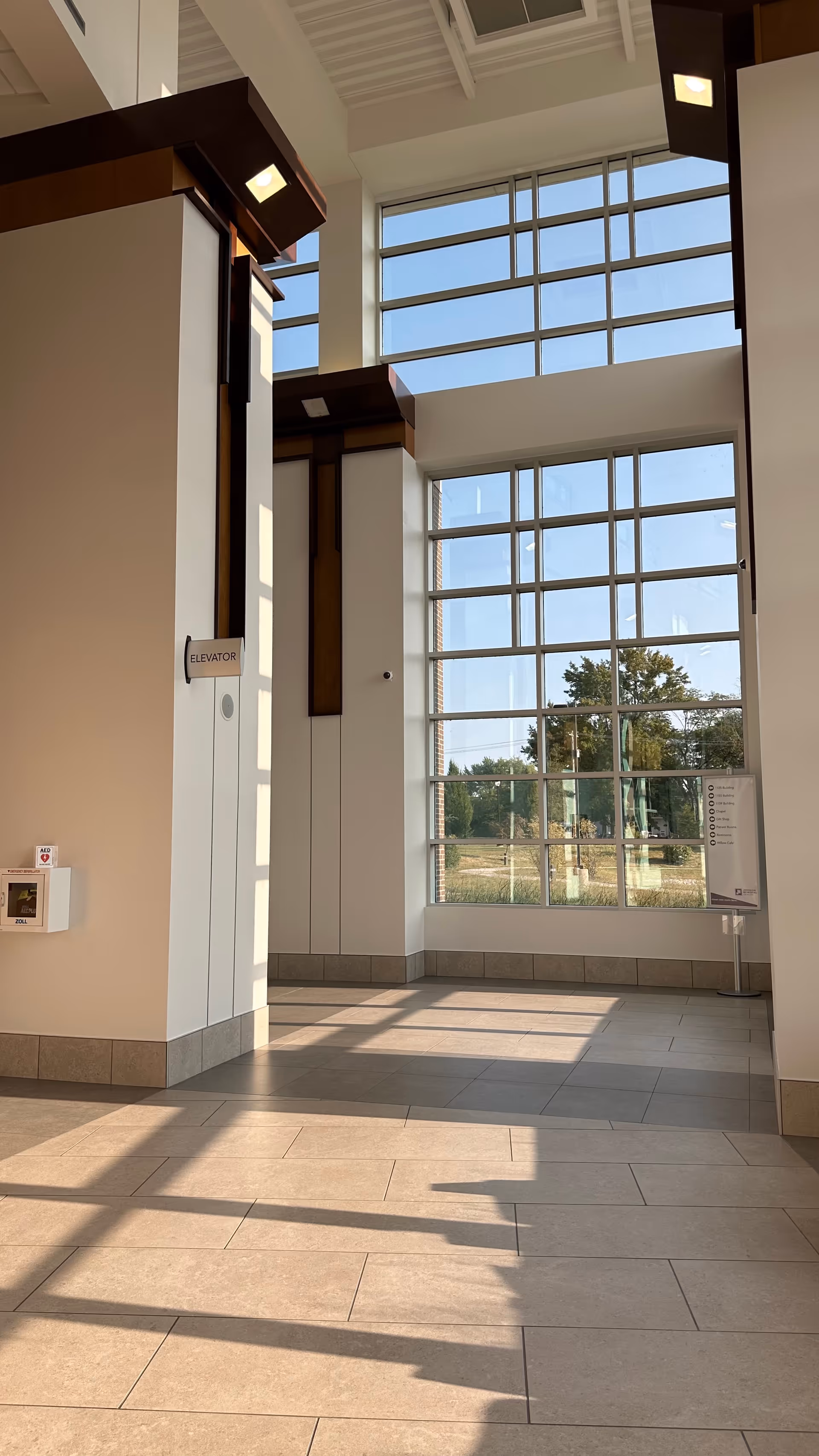 Modern hospital lobby with tall grid windows, elevator doors, and light tiled flooring.