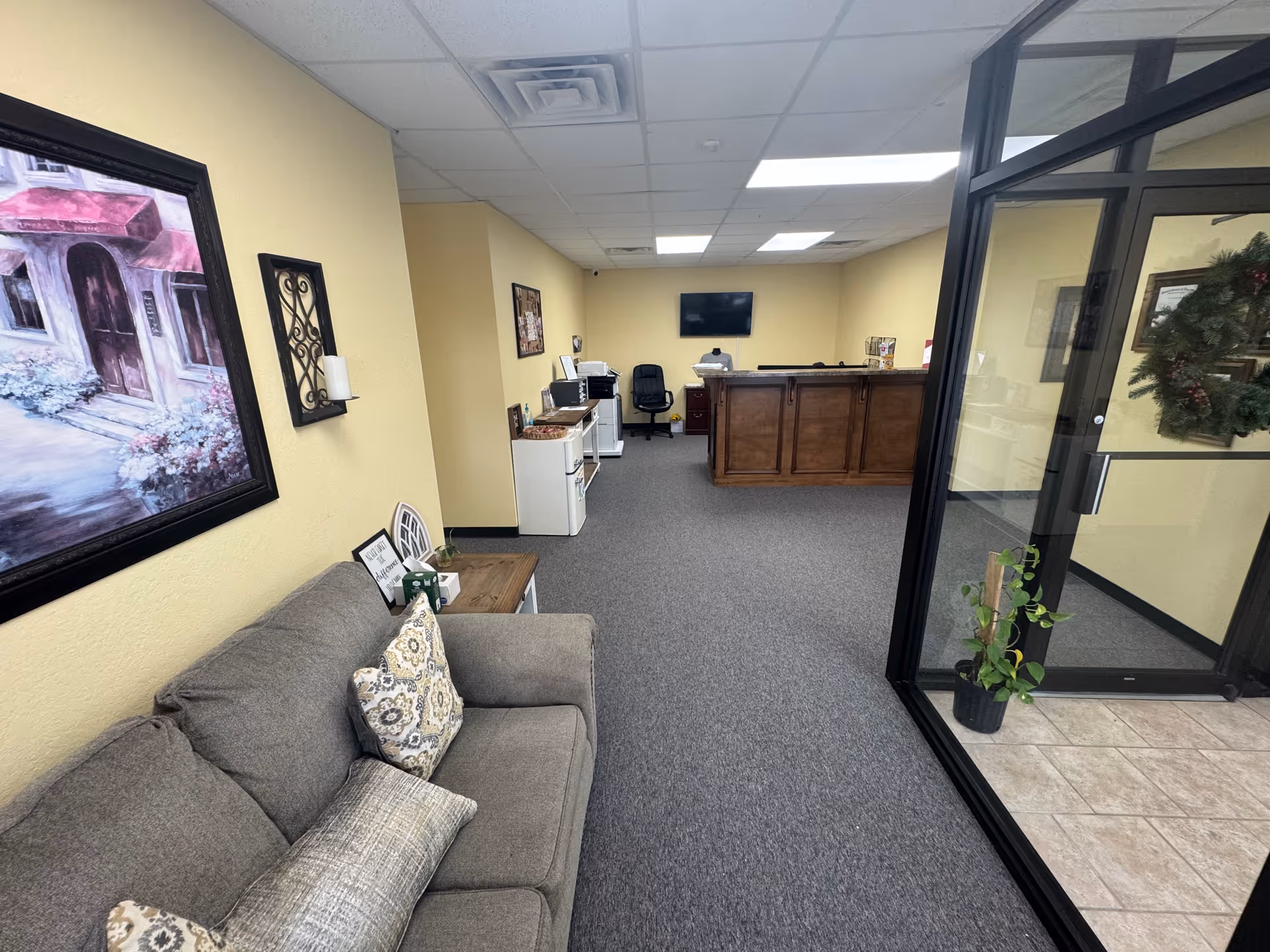 Reception area with gray couch, glass partition, and wooden front desk in a senior living facility.