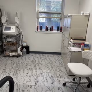 A small medical treatment room with a cart, cabinet, and rolling chair near a window.