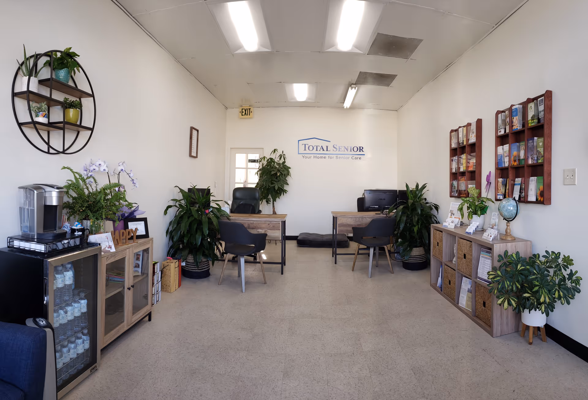 Reception lobby with desks, chairs, plants, shelves, and a Total Senior sign on the wall.