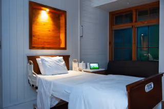 A modern hospital-style bedroom with a single bed, nightstand, and small sofa by a window.