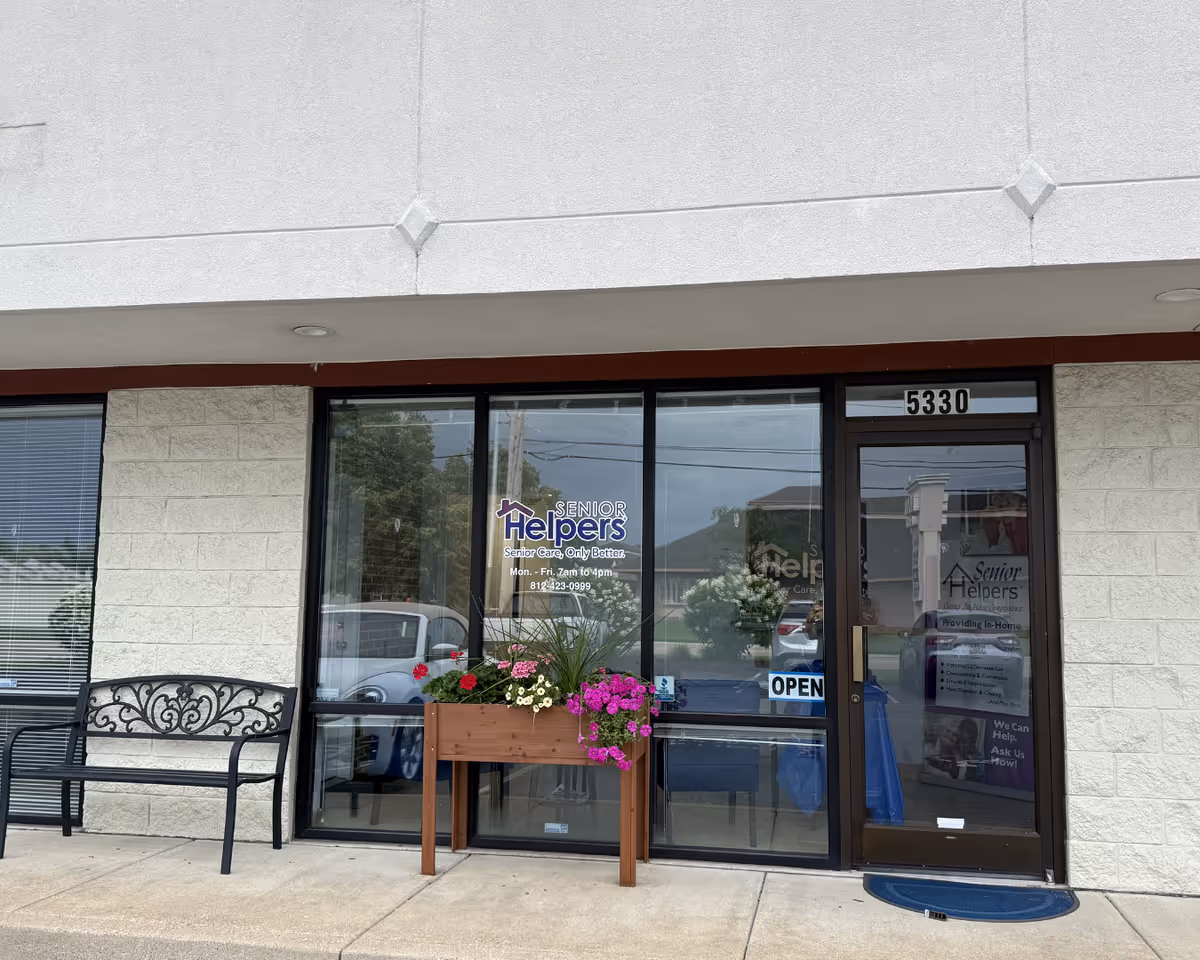 Exterior Senior Helpers storefront entrance with glass doors, a bench, and a flowering planter.