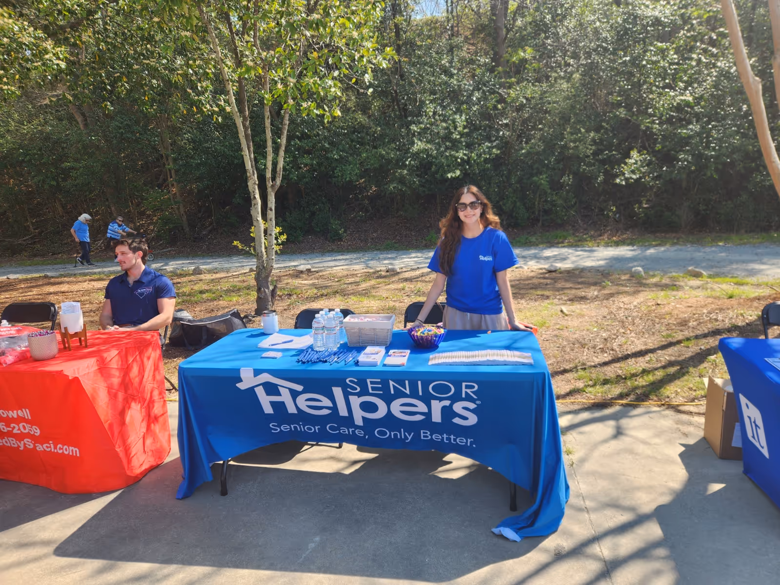 Outdoor event with a blue Senior Helpers table, staff member, and trees in the background.