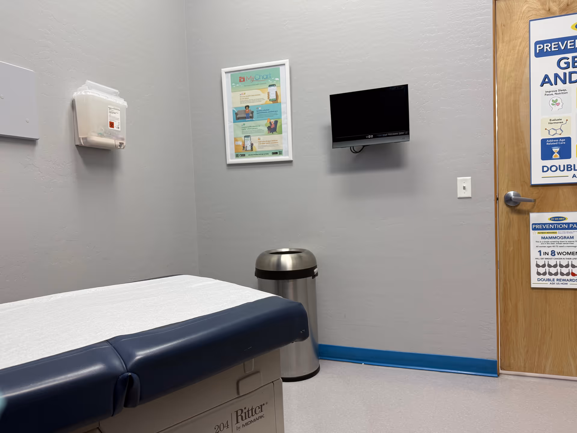 A medical examination room with a blue cushioned bed, wall TV, framed poster, and trash can.