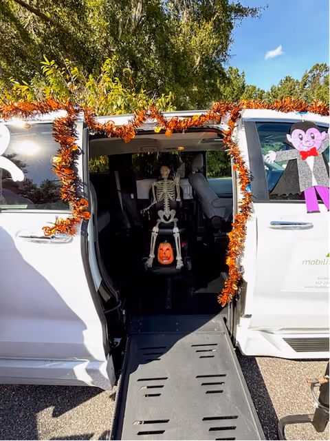 A white mobility van decorated with orange garland; a skeleton sits inside.