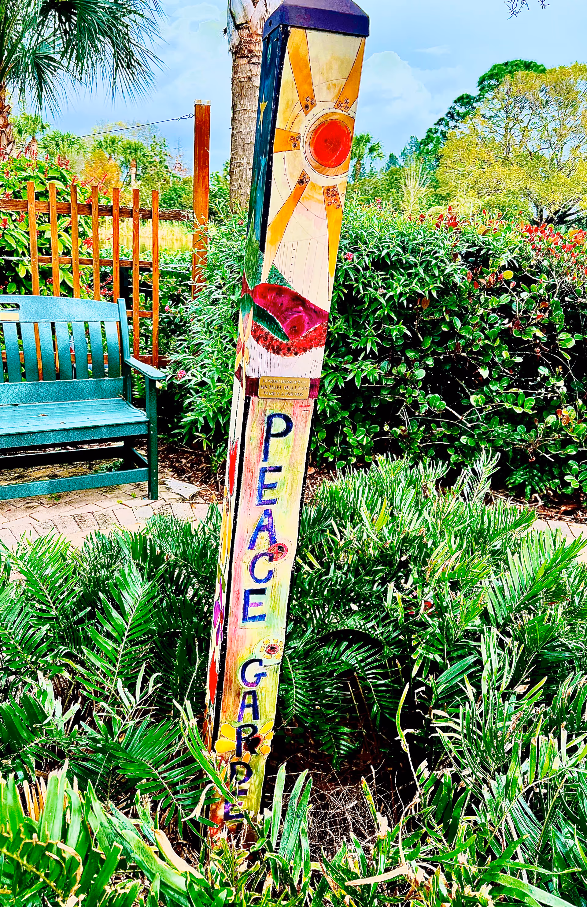 Colorful garden totem with Peace Garden text beside a green bench outdoors.