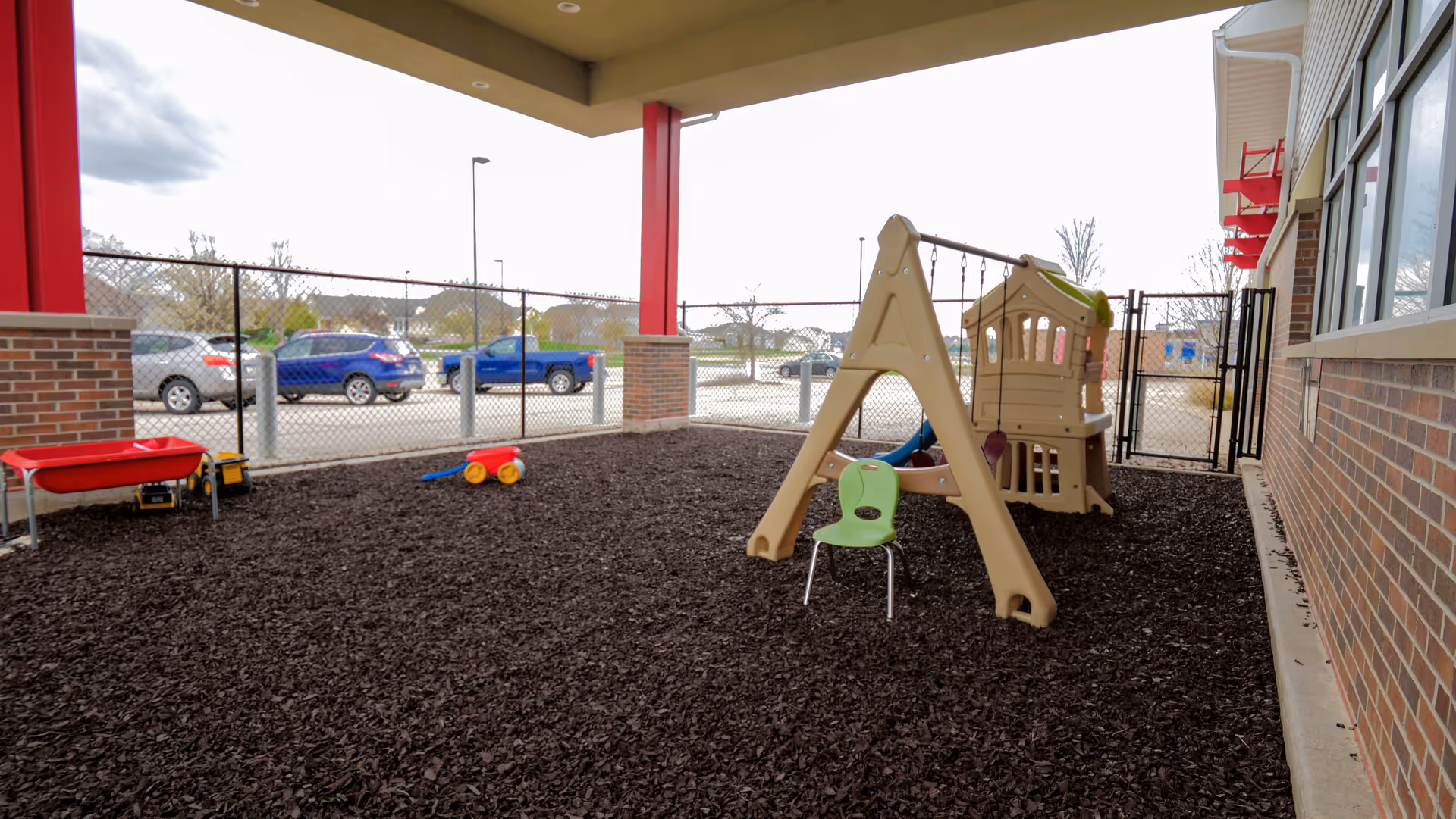 Outdoor playground area with plastic playset, mulch ground, and fence beside a building.