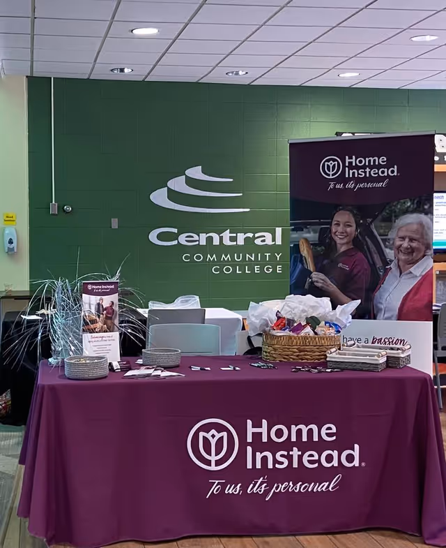 Home Instead information booth with purple tablecloth and banner indoors near a green wall.