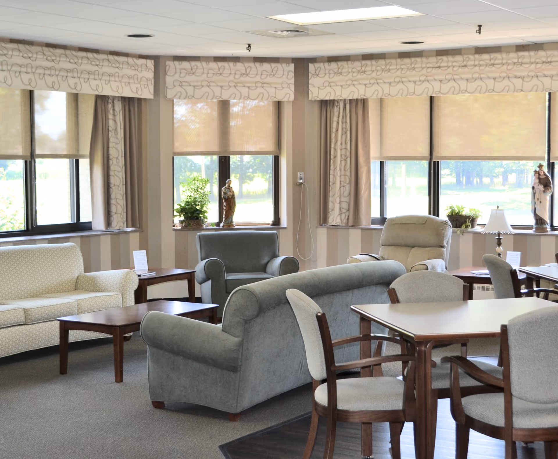Senior living lounge with beige furniture, tables, and large windows.