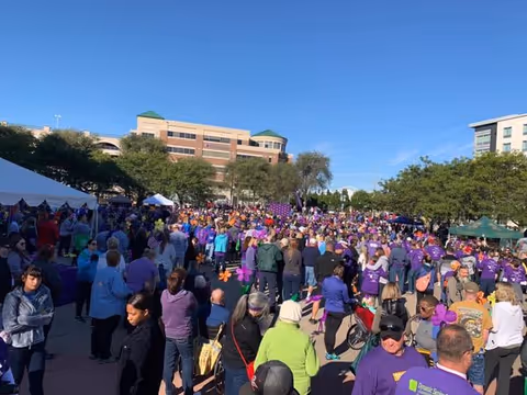 Large outdoor gathering of people in purple attire at a campus-style senior living event.