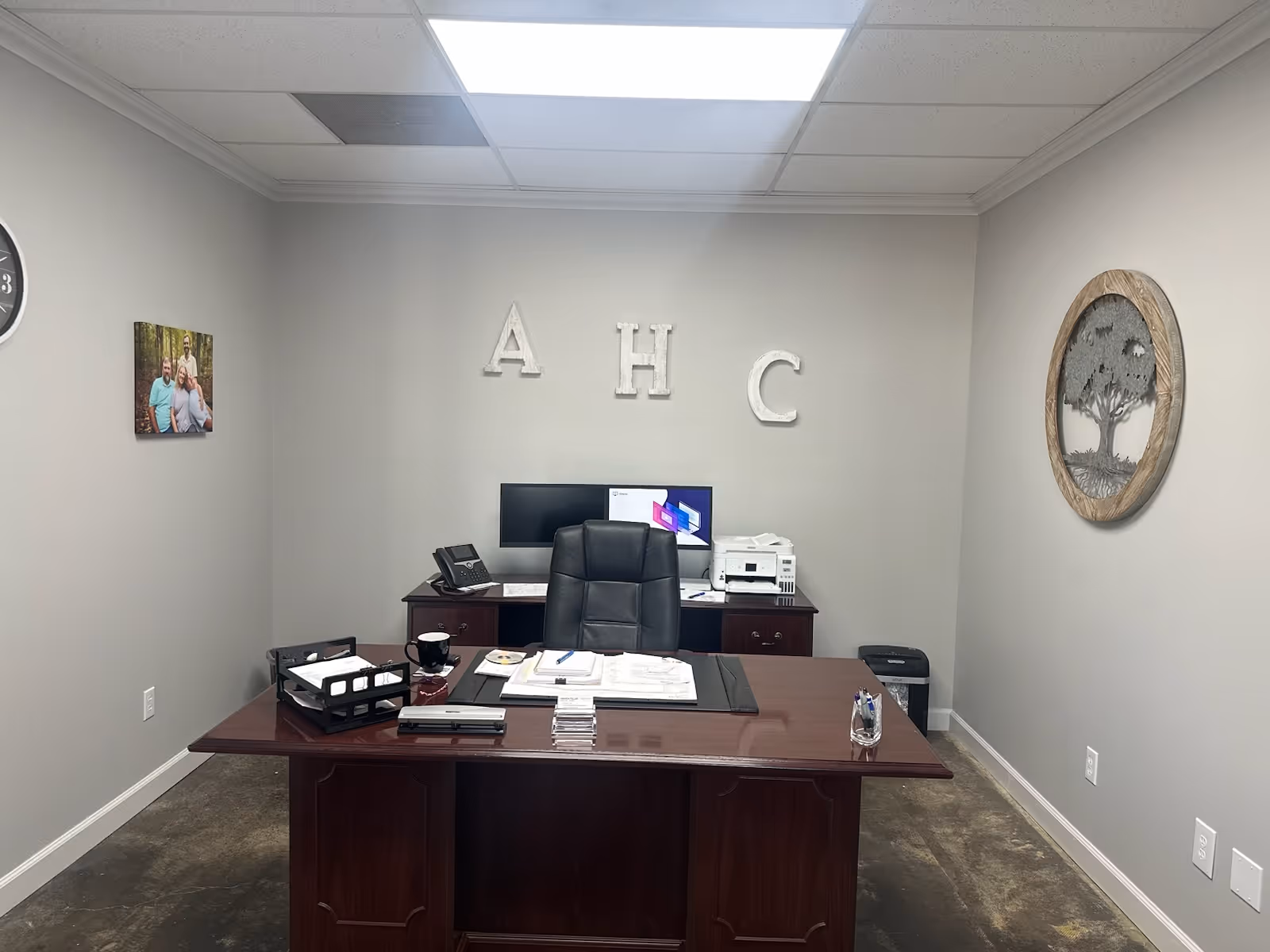Office space with a dark wooden desk, chair, computers, and AHC letters on the wall.