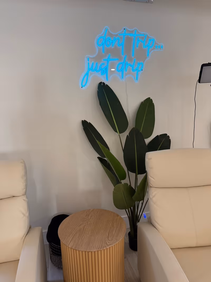 Neon blue sign reading don't trip... just-drip above a potted plant beside beige lounge chairs.