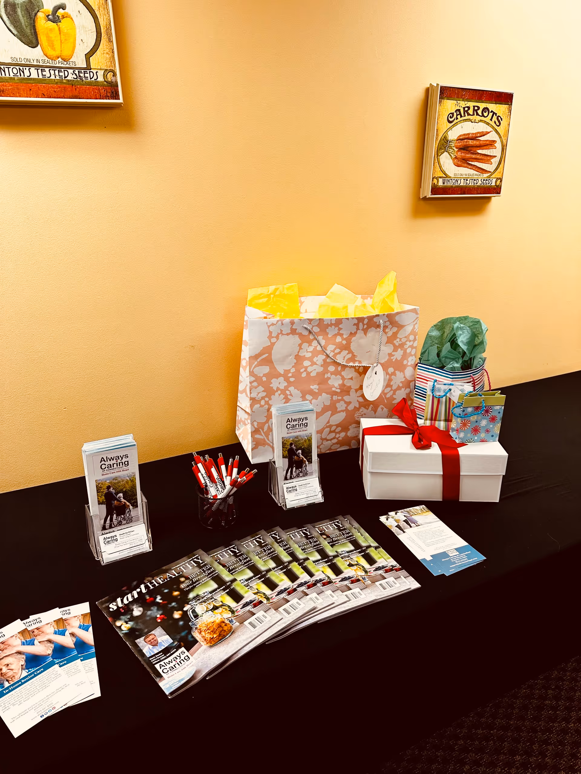 Promotional table featuring Always Caring materials, gifts, and pens against an orange wall.