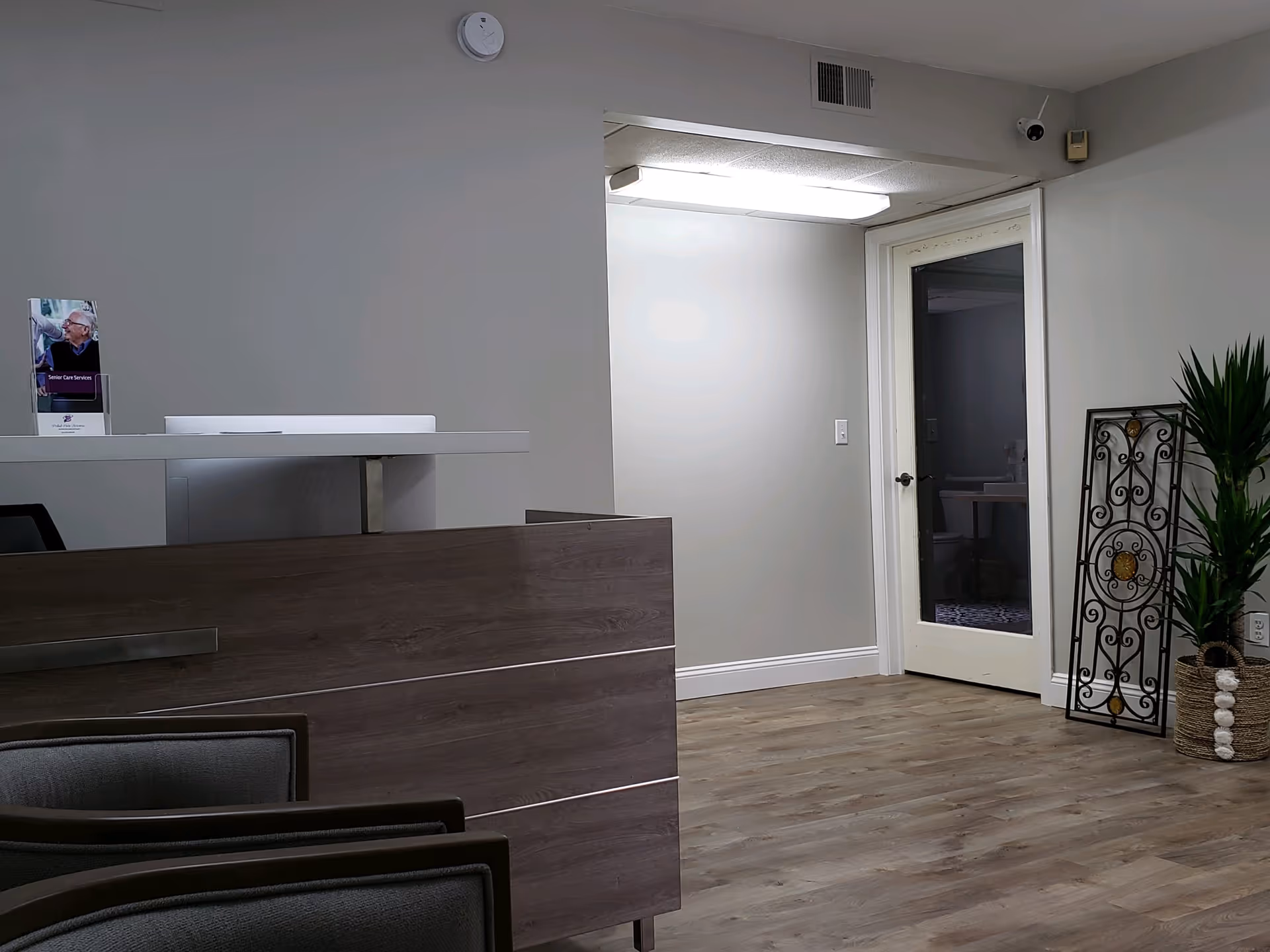 Reception desk inside a senior living facility, with gray walls, a door, and a decorative plant.