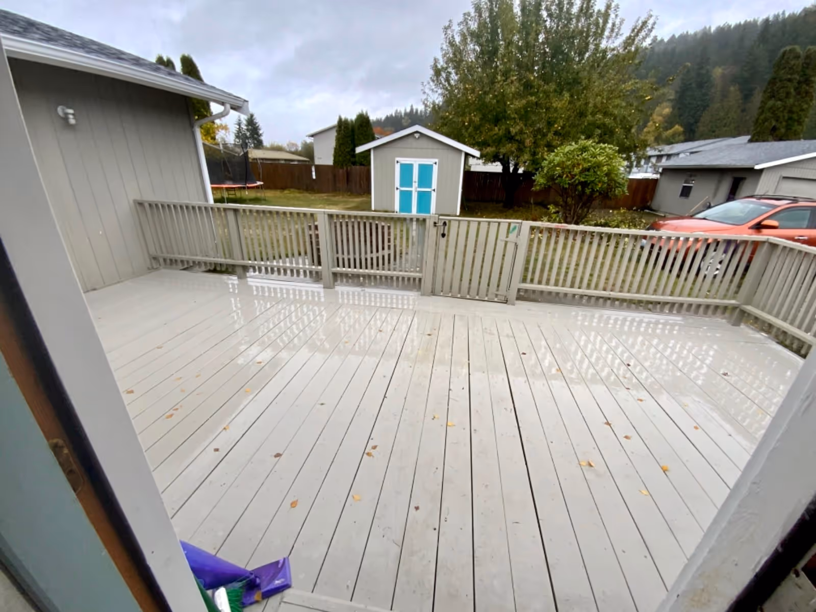 View from a back deck showing a fenced yard, shed, trees, and a parked car.