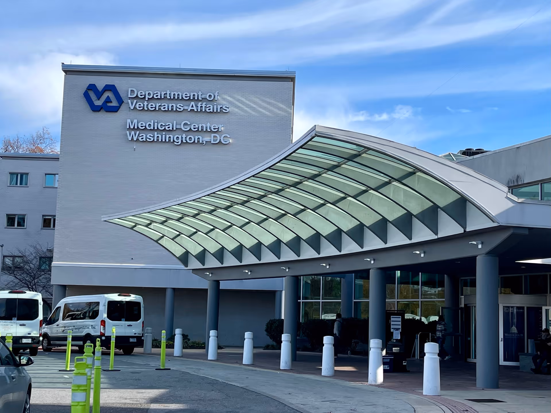 Facade of a VA Medical Center with glass canopy and entrance near parked vans.