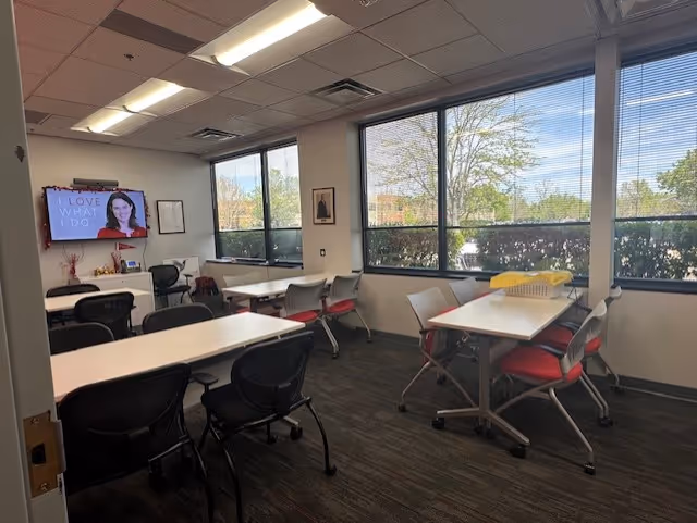 Office-style meeting room with tables, chairs, large windows, and a wall TV.