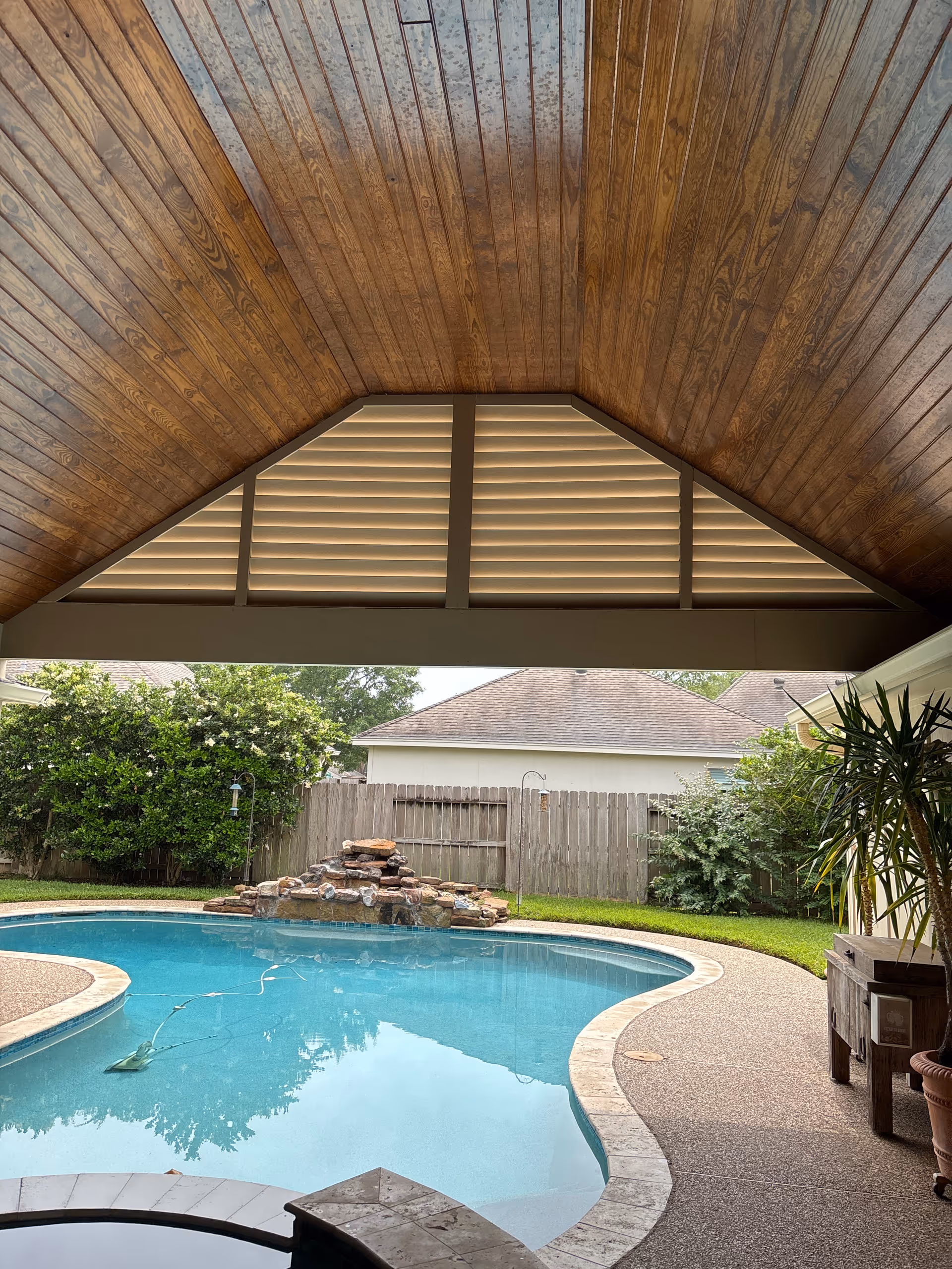 Under a wooden patio ceiling, a backyard pool with a rock waterfall and fence.