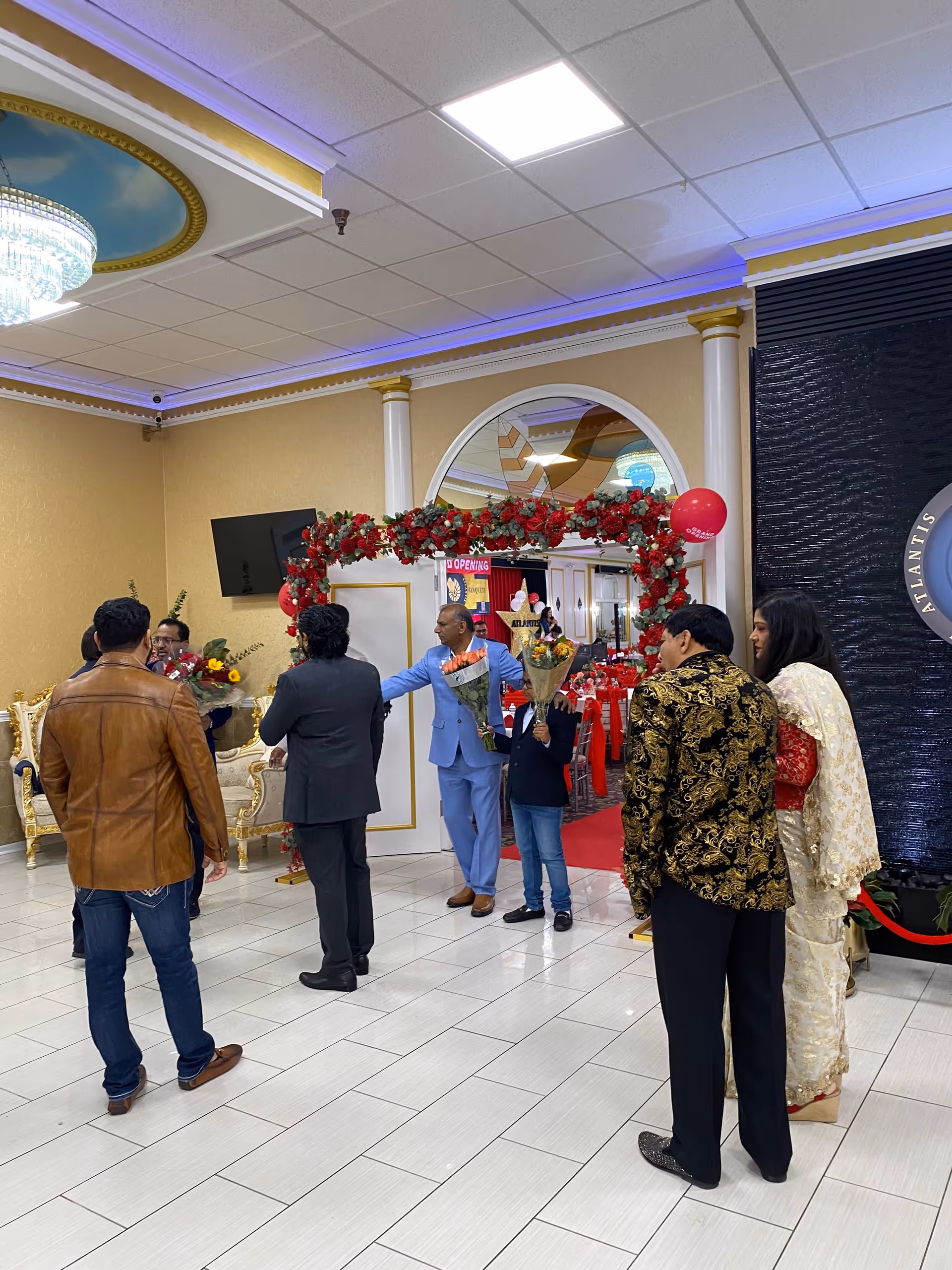 Well-dressed attendees stand in a decorated lobby with a red arch doorway and flower bouquets.