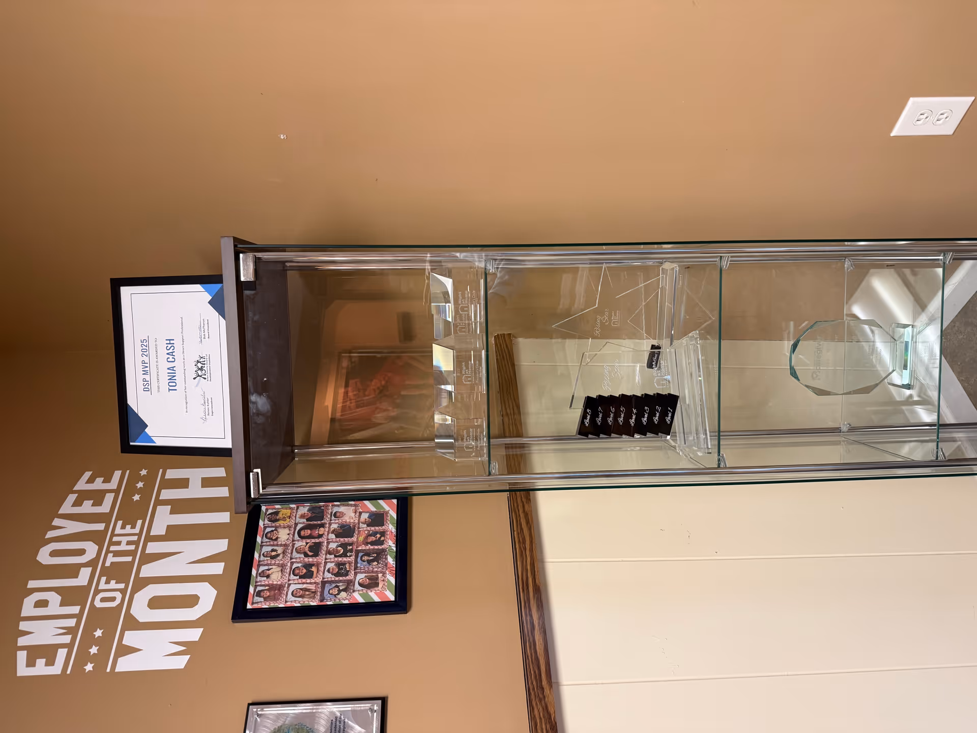 Glass display cabinet against tan wall with framed certificates and Employee of the Month sign.