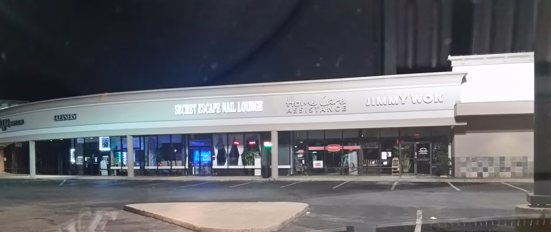 Nighttime exterior of a strip mall with multiple storefronts and a parking lot.