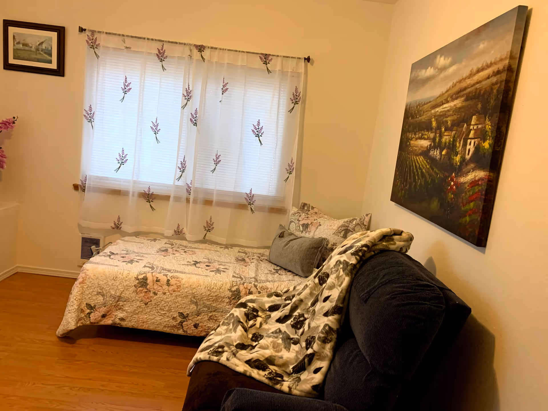 A small bedroom with a floral quilted bed, sheer curtains, wall art, and a sofa.