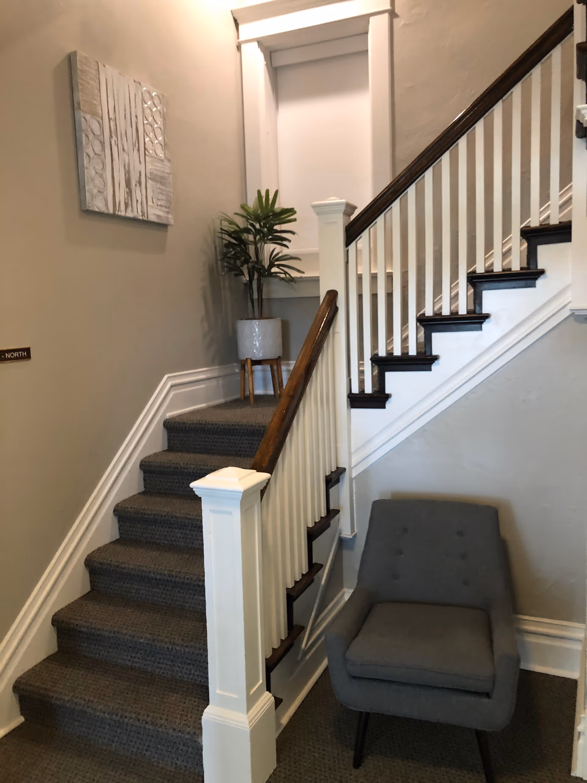 Indoor staircase with white railings, gray carpet, a gray chair, and wall art.