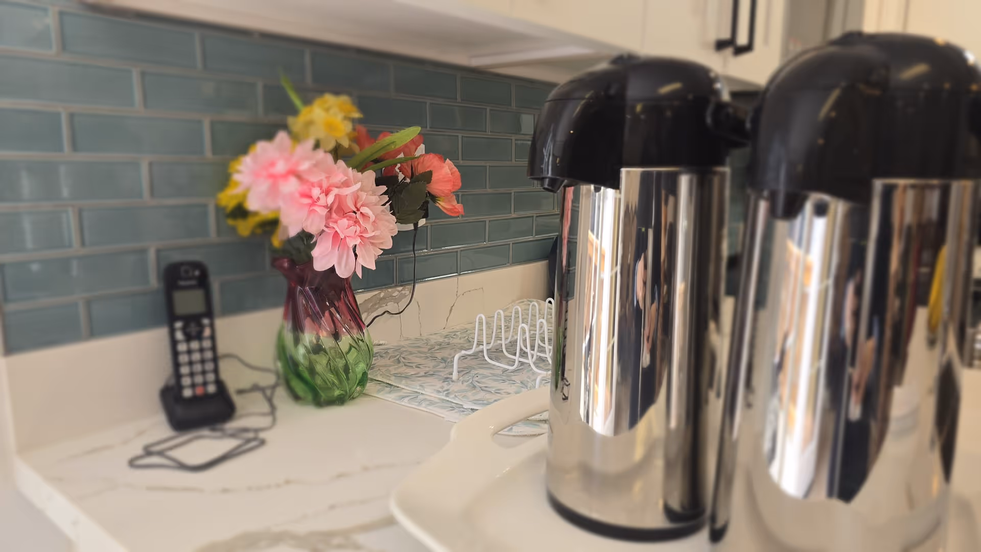 A kitchen counter with coffee urns, a phone, and flowers beside teal tile backsplash.