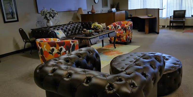 A cozy senior living lounge with tufted leather ottomans, colorful chairs, and a front desk.