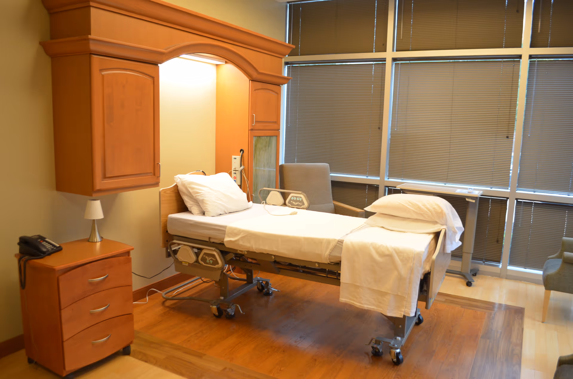 Hospital-style patient bed in a furnished private room with wood cabinets and blinds.