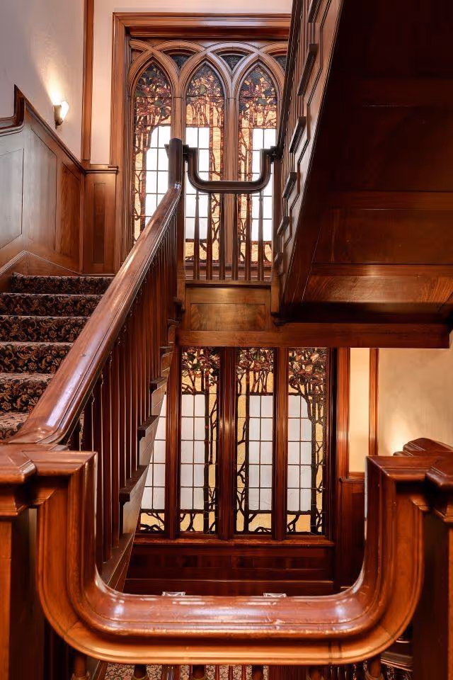 Interior wooden staircase with ornate railing and stained glass windows.