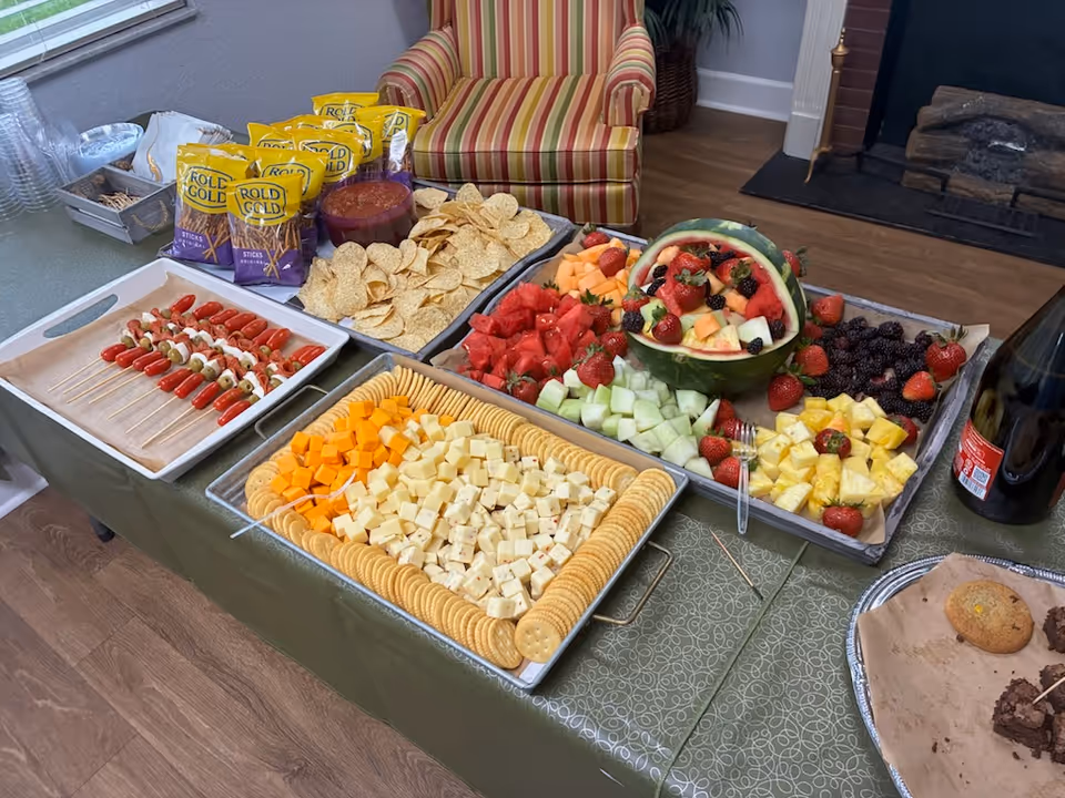 Buffet-style snack spread with chips, fruit, cheese, and skewers on a table in a living room