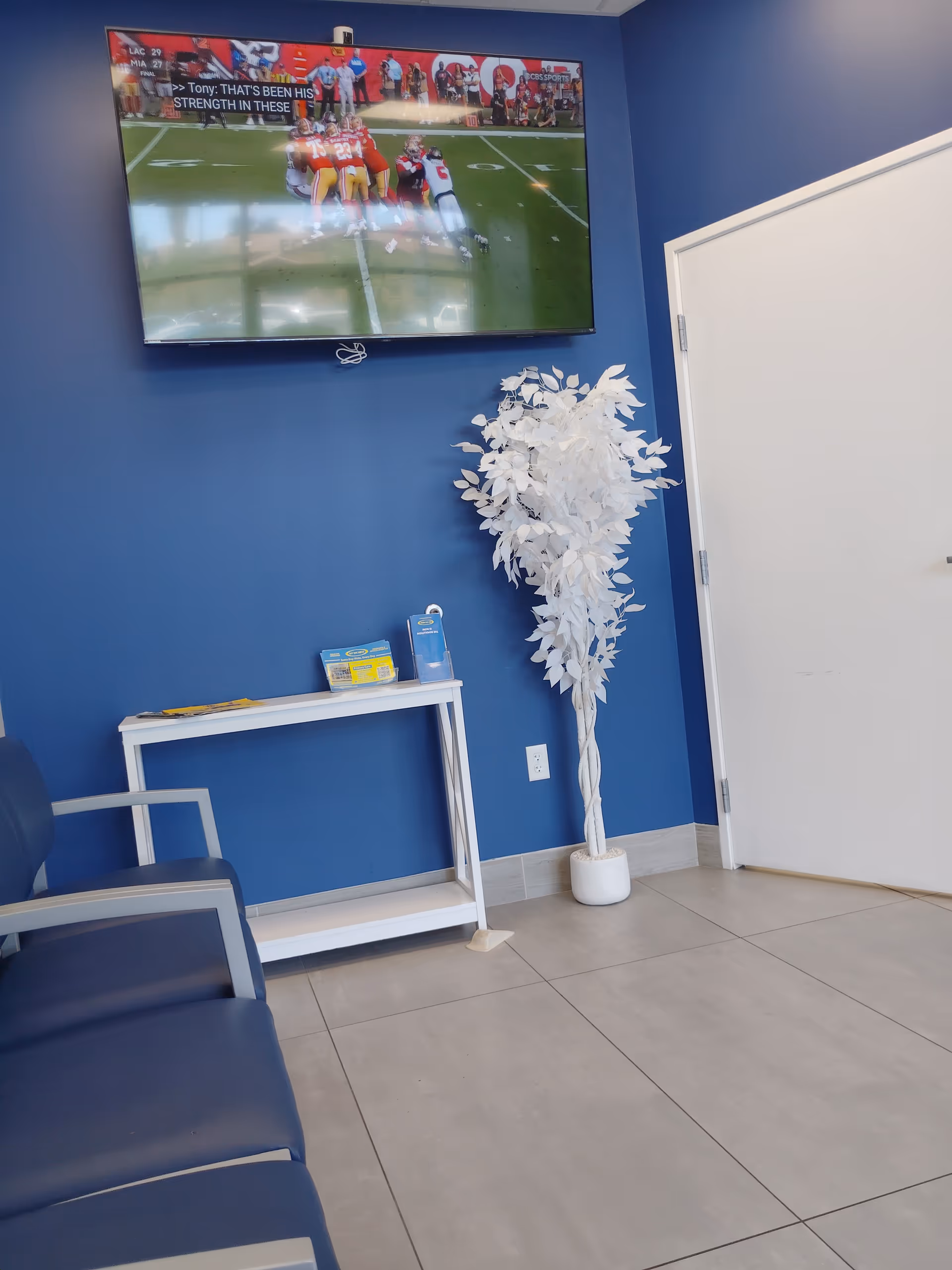 Blue-walled waiting area with wall-mounted TV, white table, brochures, and a decorative white tree.