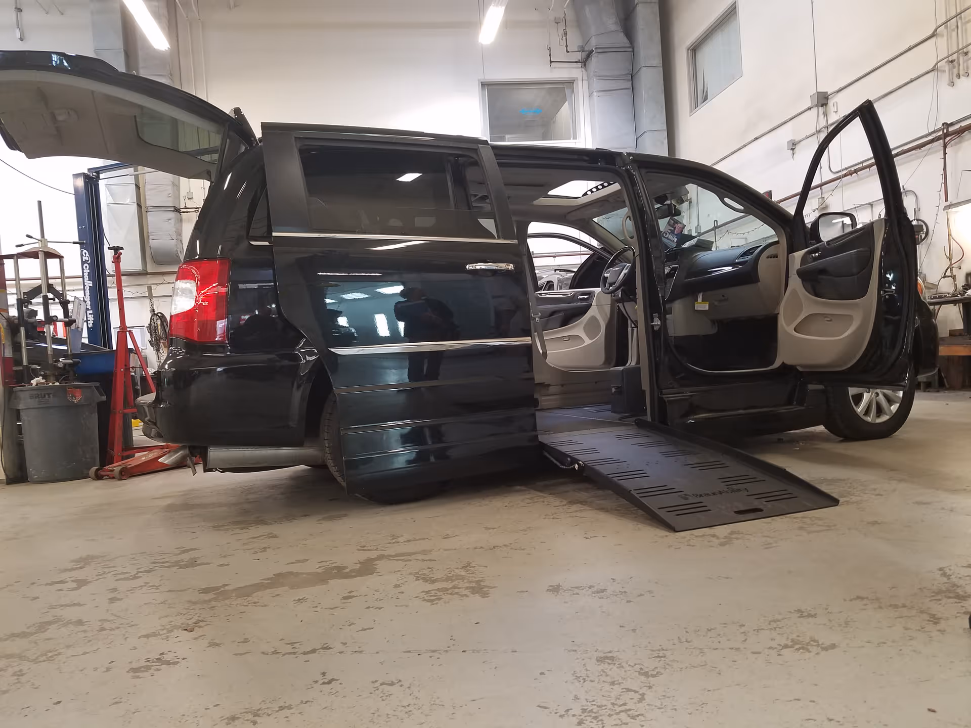 Inside a workshop, a black wheelchair-accessible van sits with its ramp extended and doors open.
