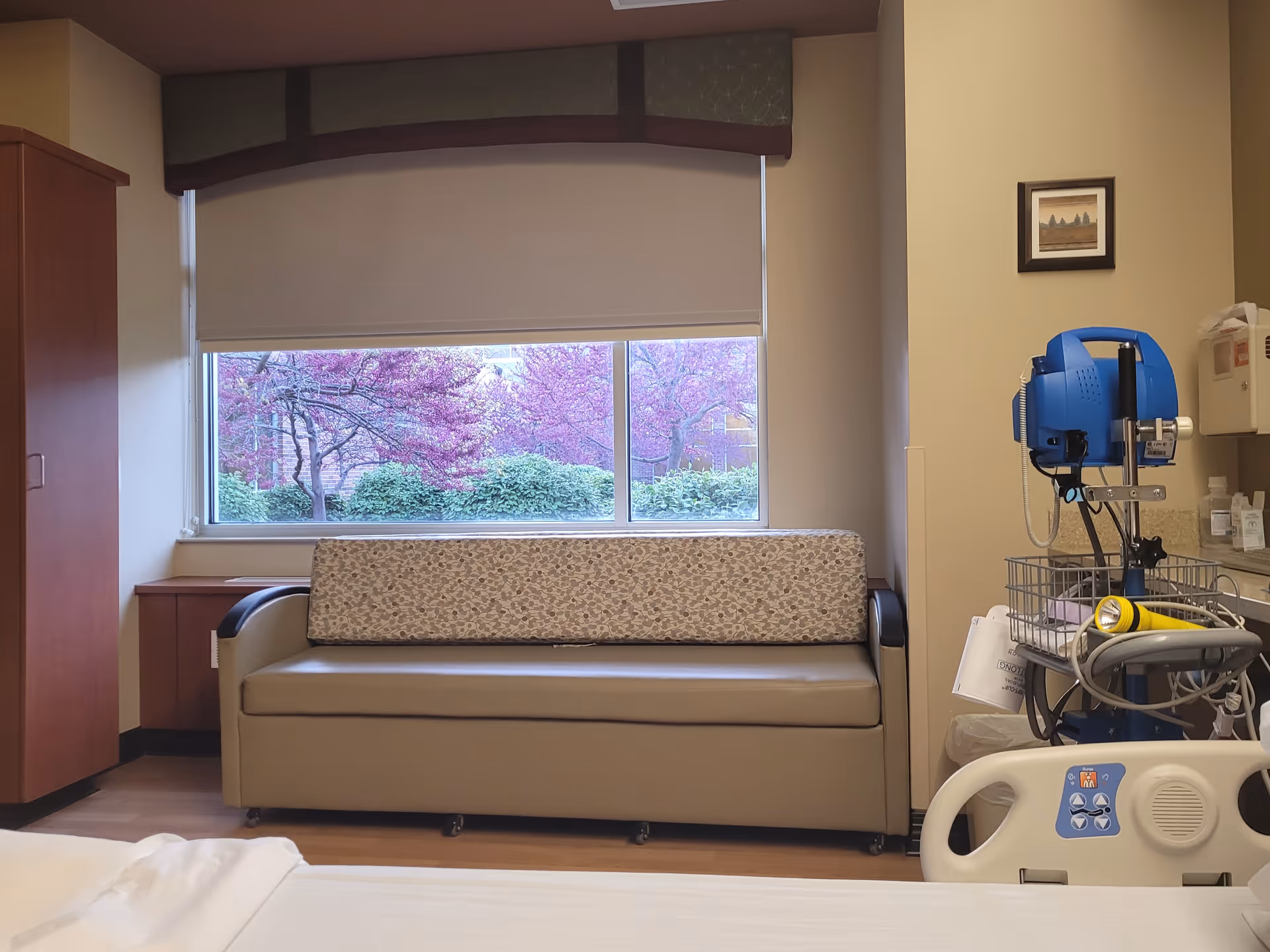 A patient room features a bed, medical cart, couch, and window view.