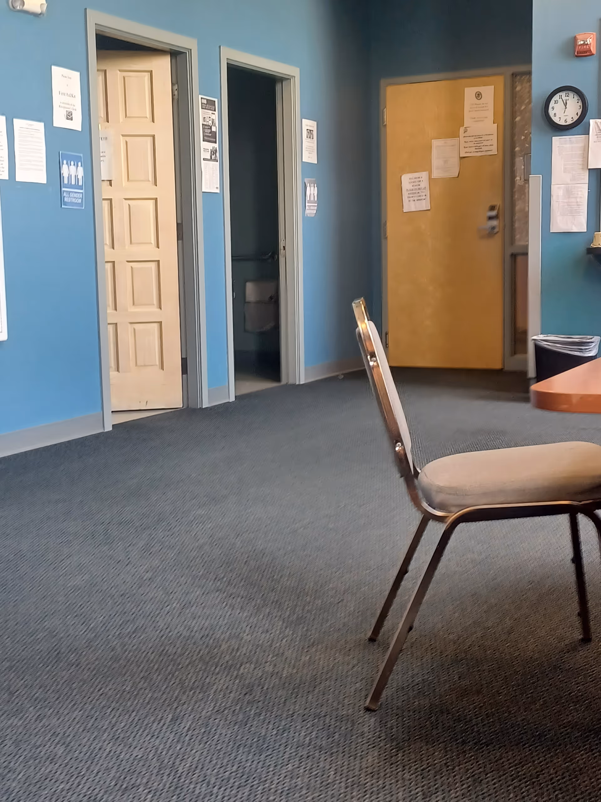 Blue-walled indoor hallway of a senior facility with open doors, a chair, and posted notices.