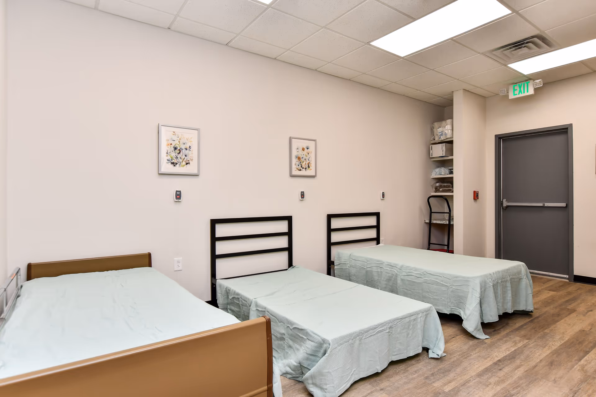 Three single beds with light blue sheets line a clean, beige-walled hospital-style bedroom.