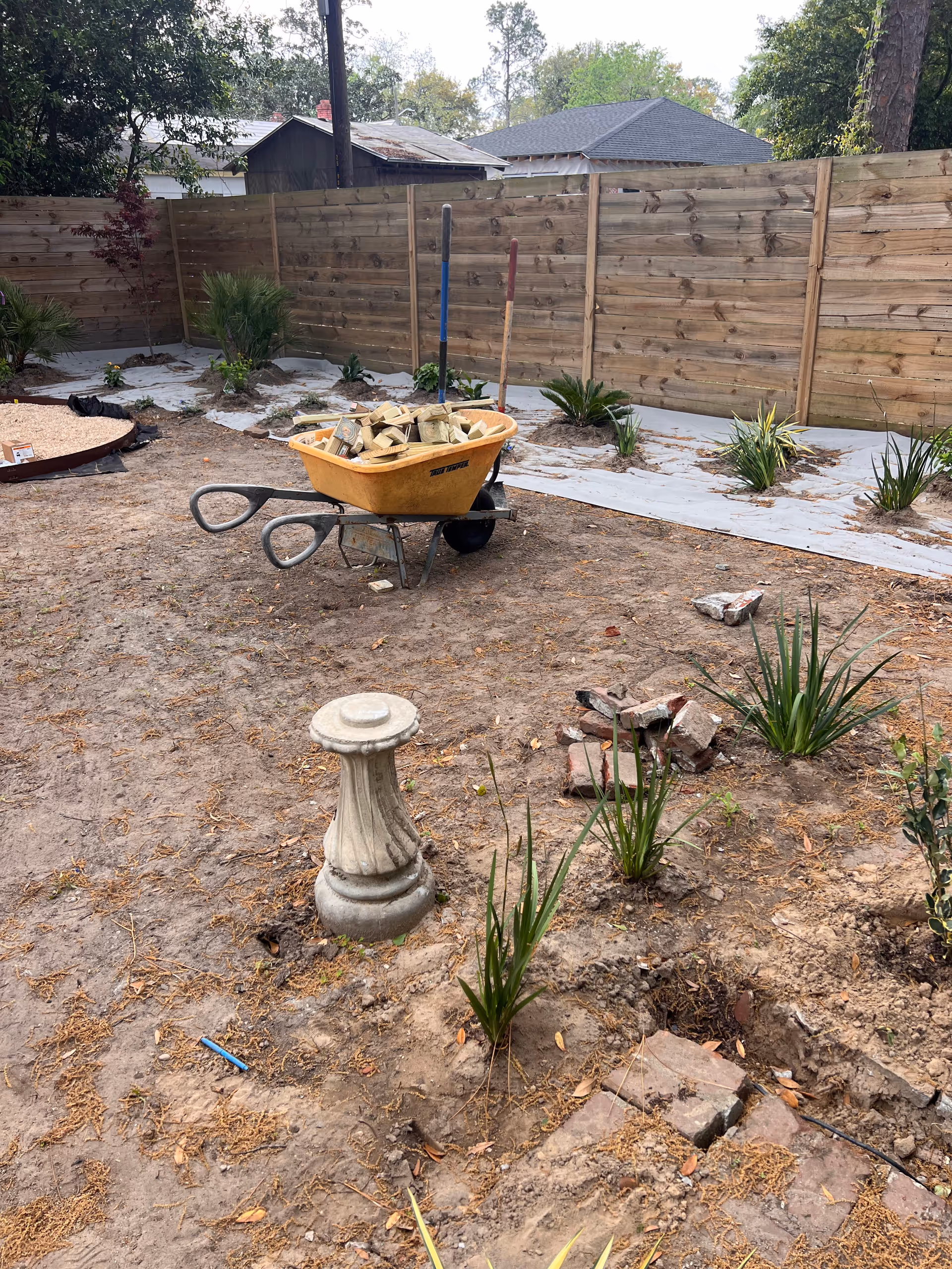 Outdoor backyard scene with a wheelbarrow of rocks, shovels, and a wooden fence.