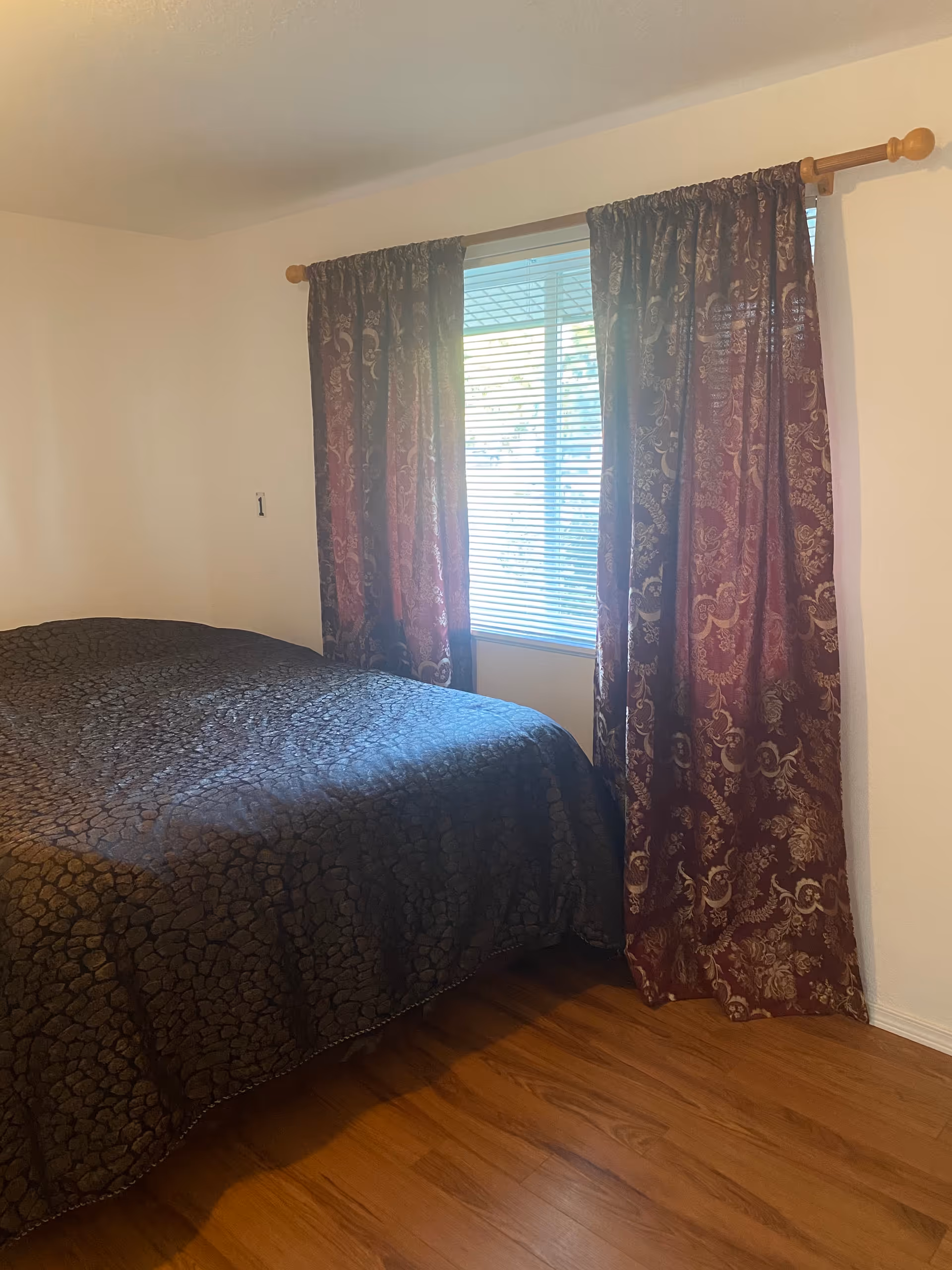 Bright bedroom with a bed, dark patterned comforter, burgundy curtains, a window, and wooden floor.