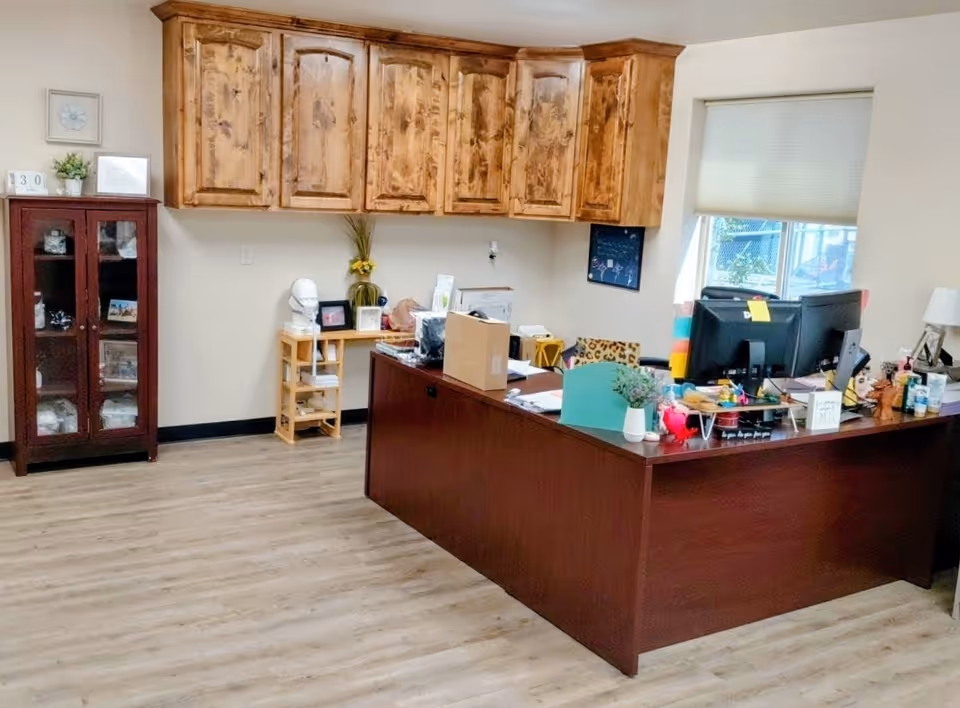 Reception area with a dark wood desk, computers, and upper cabinets in a senior living facility.